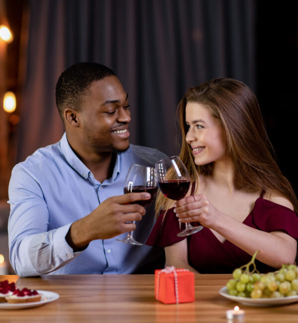 Romantic Dinner Concept. Happy Multiracial Couple Black Man And White Woman Celebrating Valentine's Day Together, Drinking Red Wine And Clinking Glasses While Dining In Restaurant, Free Space